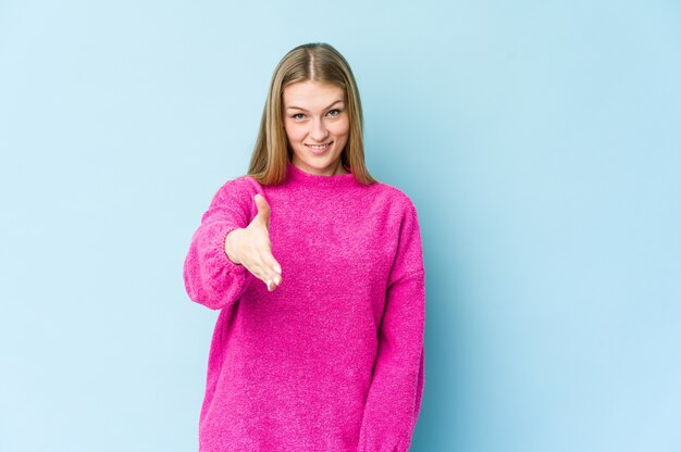 Young blonde woman isolated on blue wall stretching hand at front in greeting gesture