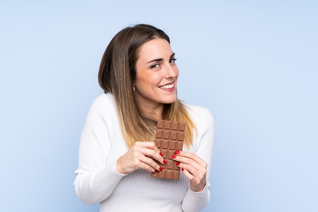 Young blonde woman over isolated blue wall taking a chocolate tablet and happy