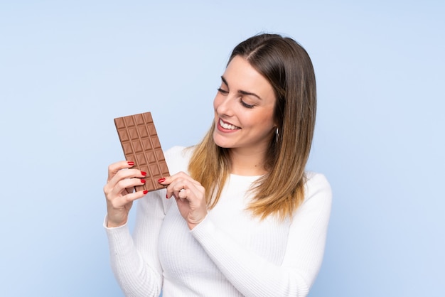 Young blonde woman over isolated blue wall taking a chocolate tablet and happy