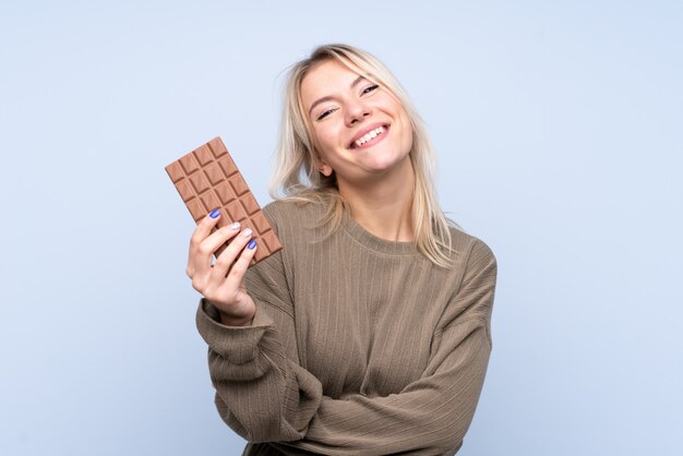 Young blonde woman over isolated blue wall taking a chocolate tablet and happy