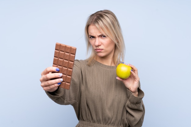 한 손에 초콜릿 태블릿을 복용하고 다른 손에 사과를 격리하는 파란색 벽 위에 젊은 금발의 여자