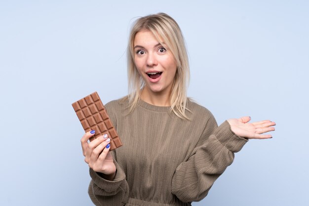 Молодая белокурая женщина над изолированной голубой стеной принимая таблетку шоколада и удивленный