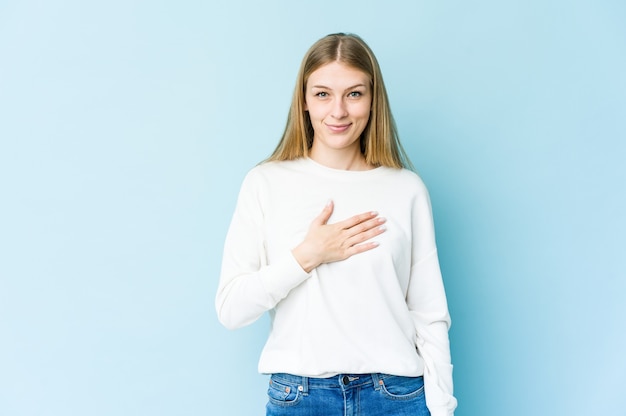 Giovane donna bionda isolata sulla parete blu che presta giuramento, mettendo la mano sul petto