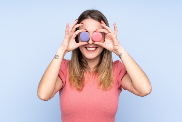 Young blonde woman over isolated blue wall wearing colorful French macarons as glasses