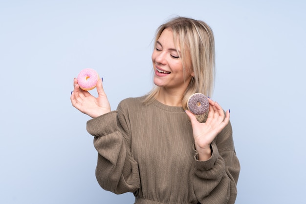 Young blonde woman over isolated blue wallholding donuts with happy expression