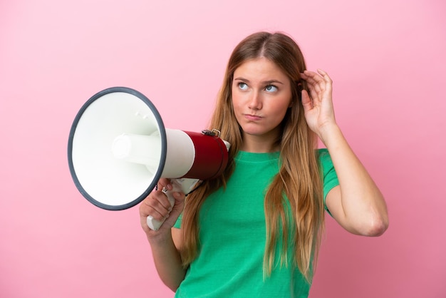 Young blonde woman isolated on pink background holding a megaphone and having doubts