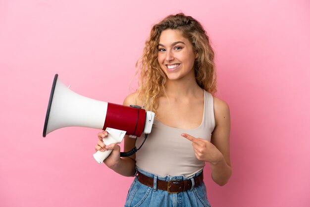 メガホンを保持し、側を指すピンクの背景に分離された若いブロンドの女性