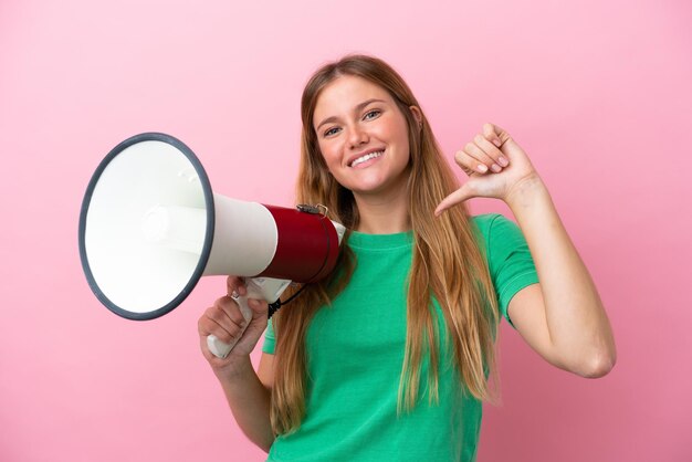 Young blonde woman isolated on pink background holding a megaphone and proud and selfsatisfied