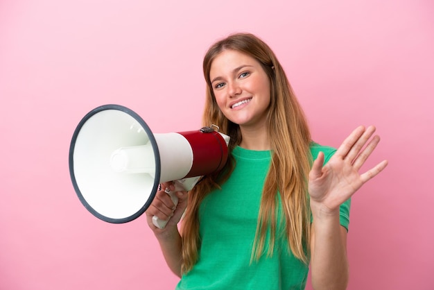 Young blonde woman isolated on pink background holding a megaphone and saluting with hand with happy expression