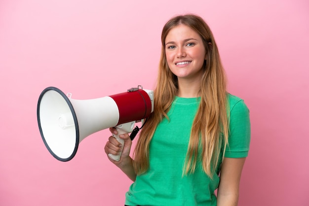 Young blonde woman isolated on pink background holding a megaphone and smiling a lot