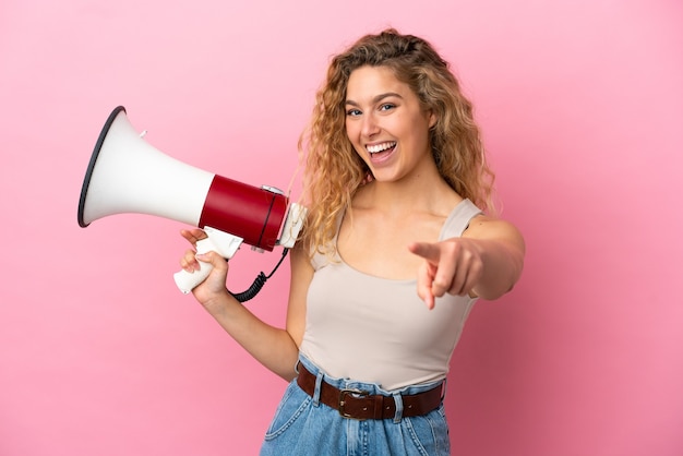 Young blonde woman isolated on pink background holding a megaphone and smiling while pointing to the front