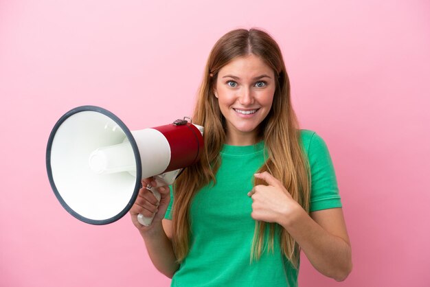 Young blonde woman isolated on pink background holding a megaphone and with surprise facial expression