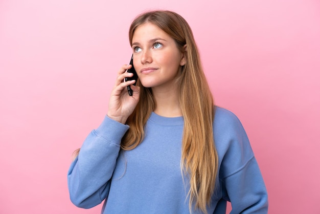 Young blonde woman isolated on pink background keeping a conversation with the mobile phone with someone