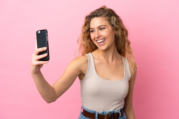 Young blonde woman isolated on pink background making a selfie with mobile phone