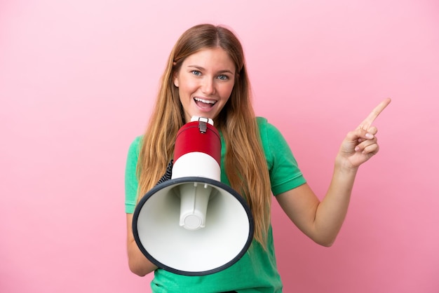 Young blonde woman isolated on pink background shouting through a megaphone and pointing side