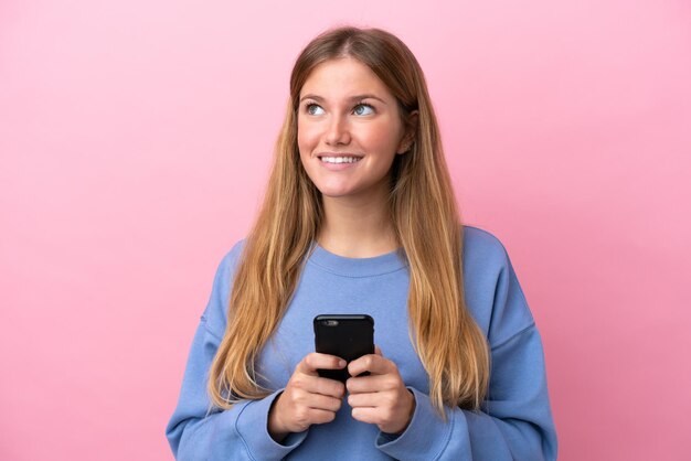 Young blonde woman isolated on pink background using mobile phone and looking up