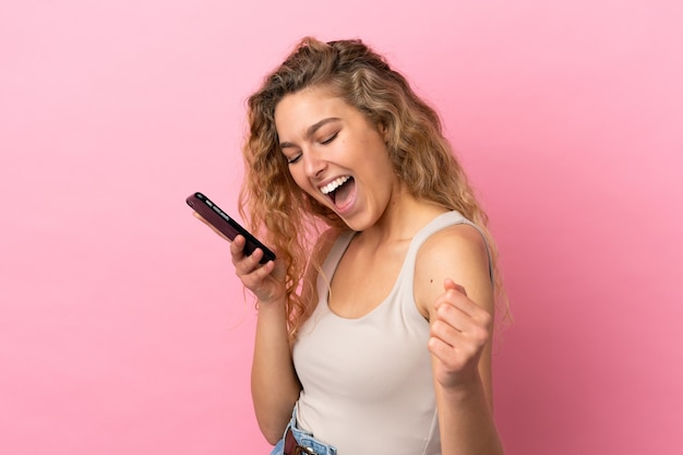 Young blonde woman isolated on pink background using mobile phone and singing