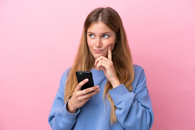 Young blonde woman isolated on pink background using mobile phone and thinking