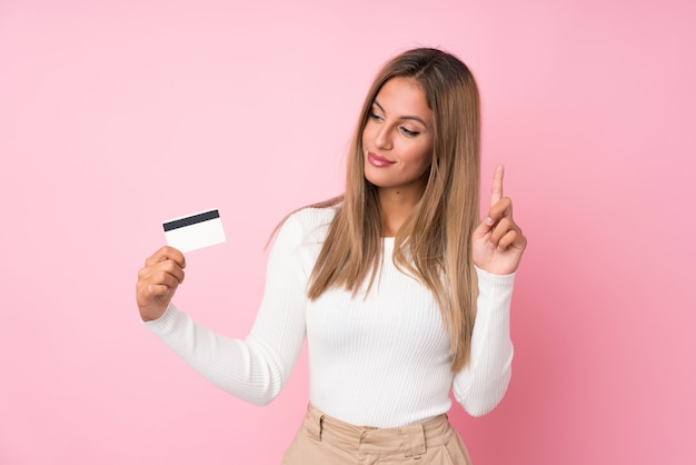 Young blonde woman over isolated pink wall holding a credit card