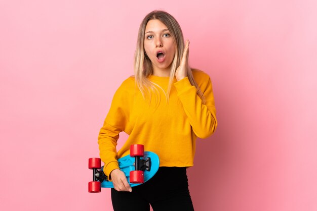 Young blonde woman isolated on pink wall with a skate and doing surprise gesture