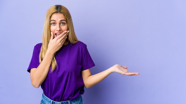 Young blonde woman isolated on purple background holds copy space on a palm, keep hand over cheek. Amazed and delighted.