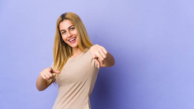 Young blonde woman isolated on purple pointing to front with fingers.