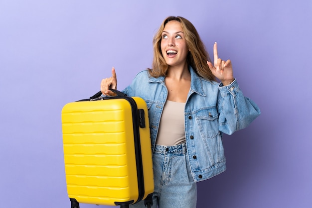 Photo young blonde woman isolated on purple in vacation with travel suitcase and pointing up