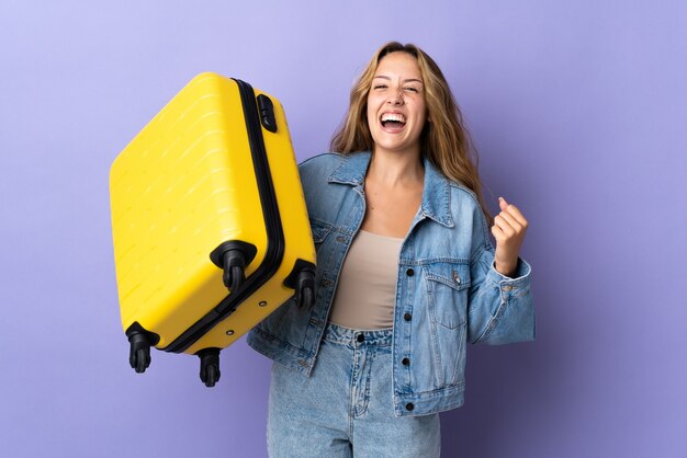 Young blonde woman isolated on purple in vacation with travel suitcase