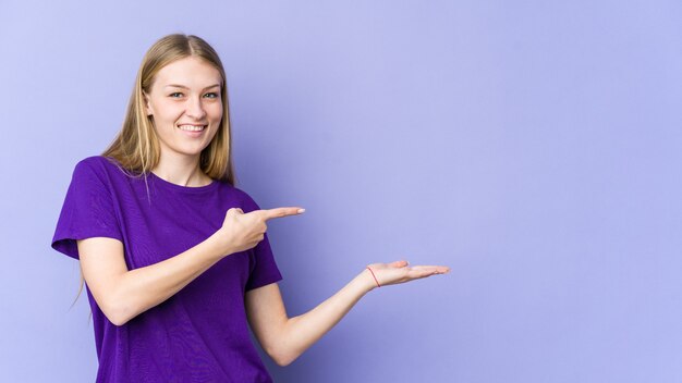 Young blonde woman isolated on purple wall excited holding a copy space on palm