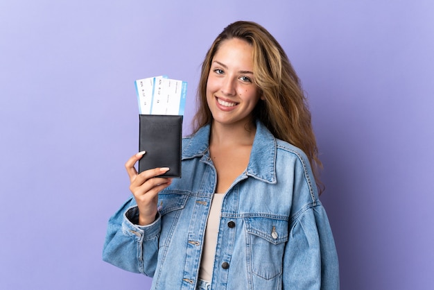 Young blonde woman isolated on purple wall happy in vacation with passport and plane tickets