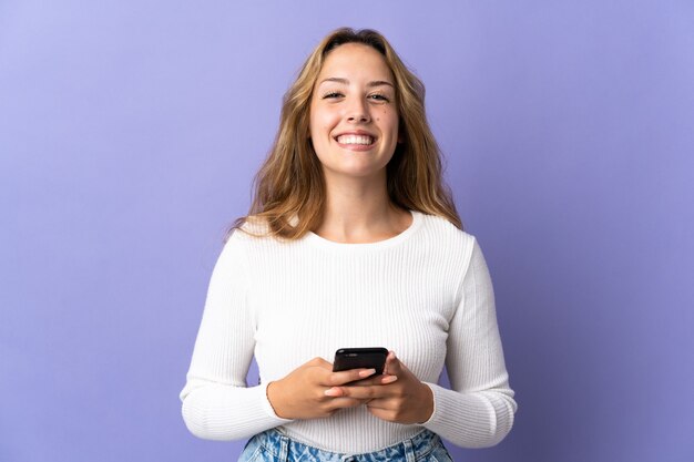 Young blonde woman isolated on purple wall looking at the camera and smiling while using the mobile