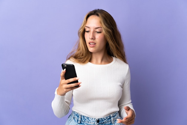 Young blonde woman isolated on purple wall sending a message or email with the mobile