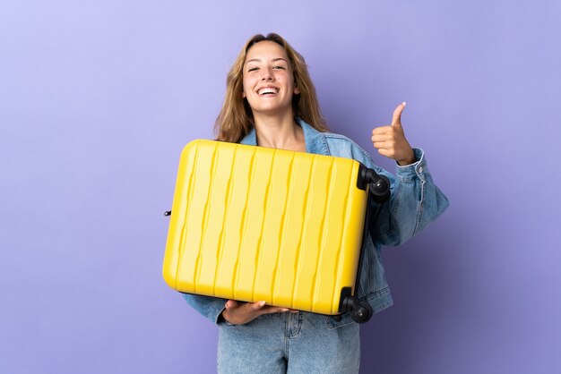 Young blonde woman isolated on purple wall in vacation with travel suitcase and with thumb up