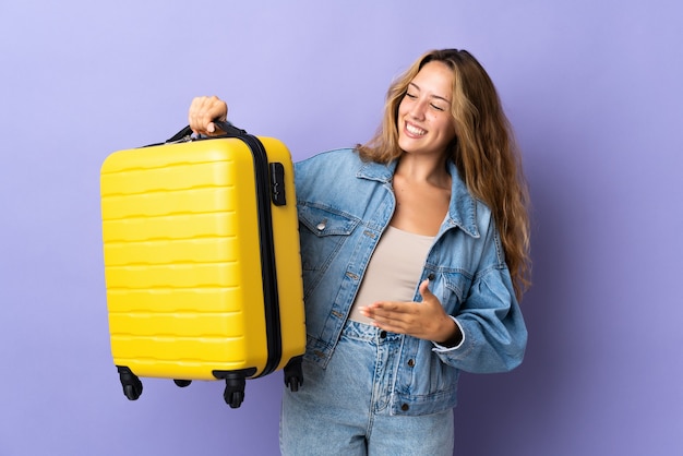 Молодая блондинка женщина изолирована на фиолетовой стене в отпуске с чемоданом путешествия