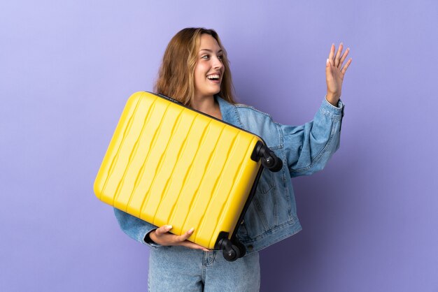 Young blonde woman isolated in vacation with travel suitcase saluting