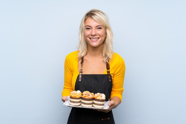 Young blonde woman over isolated wall holding mini cakes