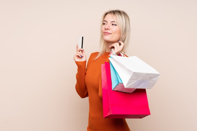 Young blonde woman over isolated wall holding shopping bags and a credit card and thinking