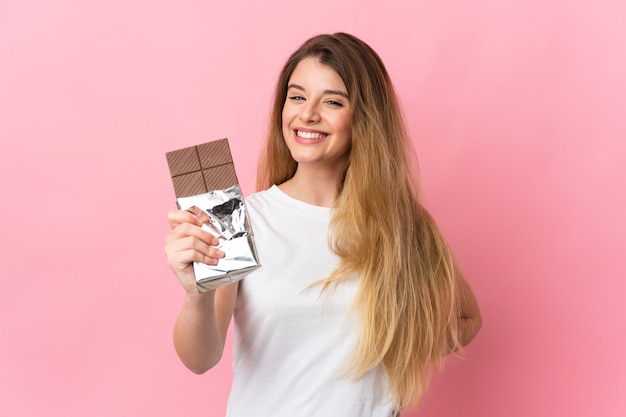 Giovane donna bionda sopra la parete isolata che prende una compressa di cioccolato e felice