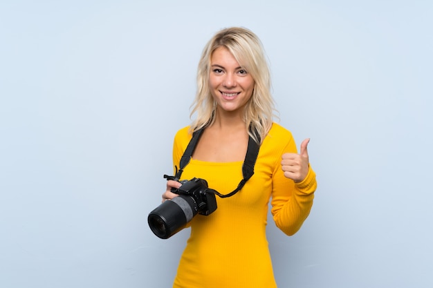 Молодая блондинка над изолированной стеной с профессиональной камерой и с большим пальцем вверх