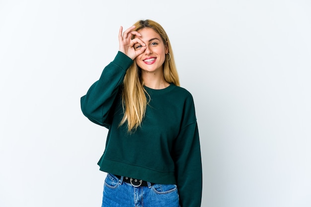 Young blonde woman isolated on white excited keeping ok gesture on eye.