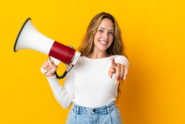 Young blonde woman isolated on yellow background holding a megaphone and smiling while pointing to the front