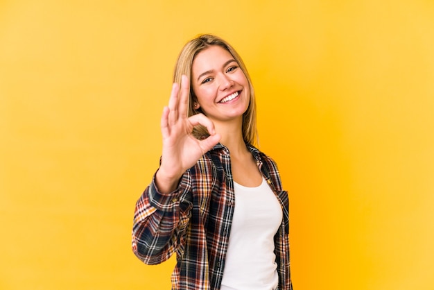 Young blonde woman isolated on a yellow wall cheerful and confident showing ok gesture