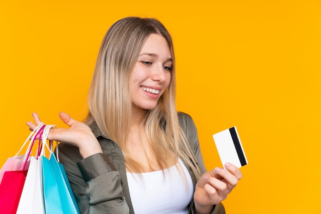 Young blonde woman over isolated yellow wall holding shopping bags and a credit card