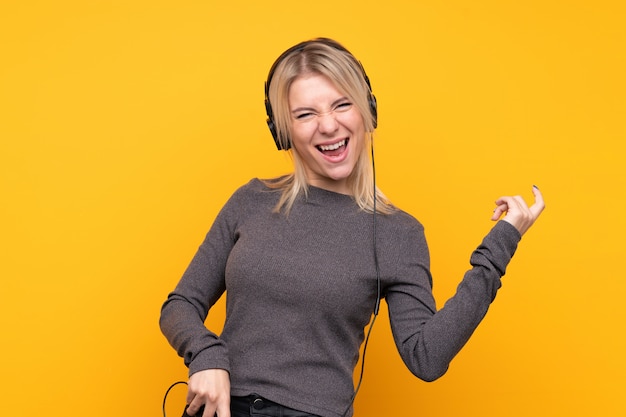 Young blonde woman over isolated yellow wall listening music and doing guitar gesture