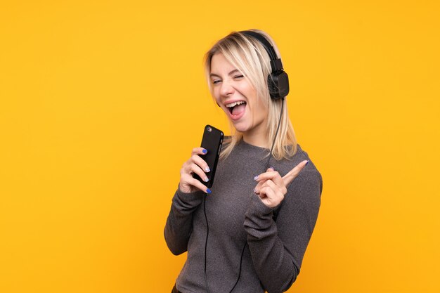 Young blonde woman over isolated yellow wall listening music with a mobile and singing