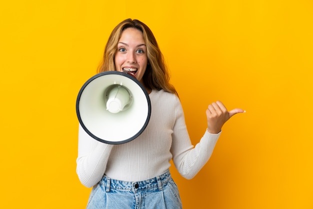 Photo young blonde woman isolated on yellow wall shouting through a megaphone and pointing side