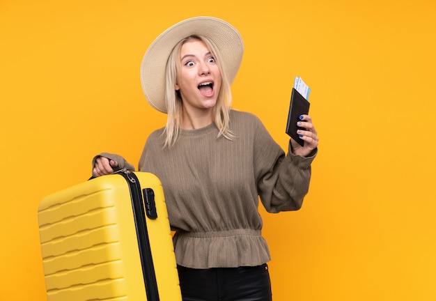 Молодая белокурая женщина над изолированной желтой стеной в каникулах с чемоданом и паспортом и удивленная