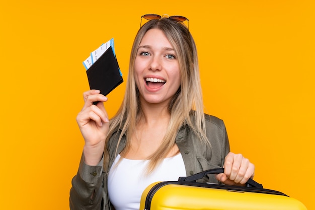 Young blonde woman over isolated yellow wall in vacation with suitcase and passport