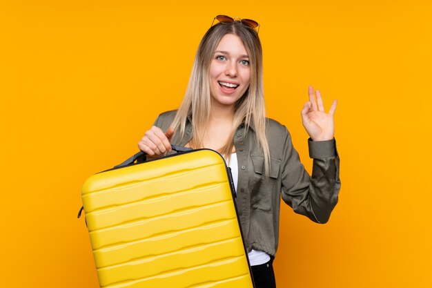 Young blonde woman over isolated yellow wall in vacation with travel suitcase and a hat