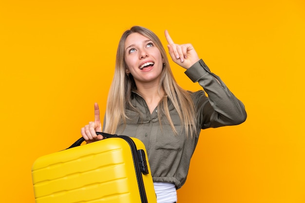 여행 가방 및 가리키는 휴가에 고립 된 노란색 벽 위에 젊은 금발의 여자